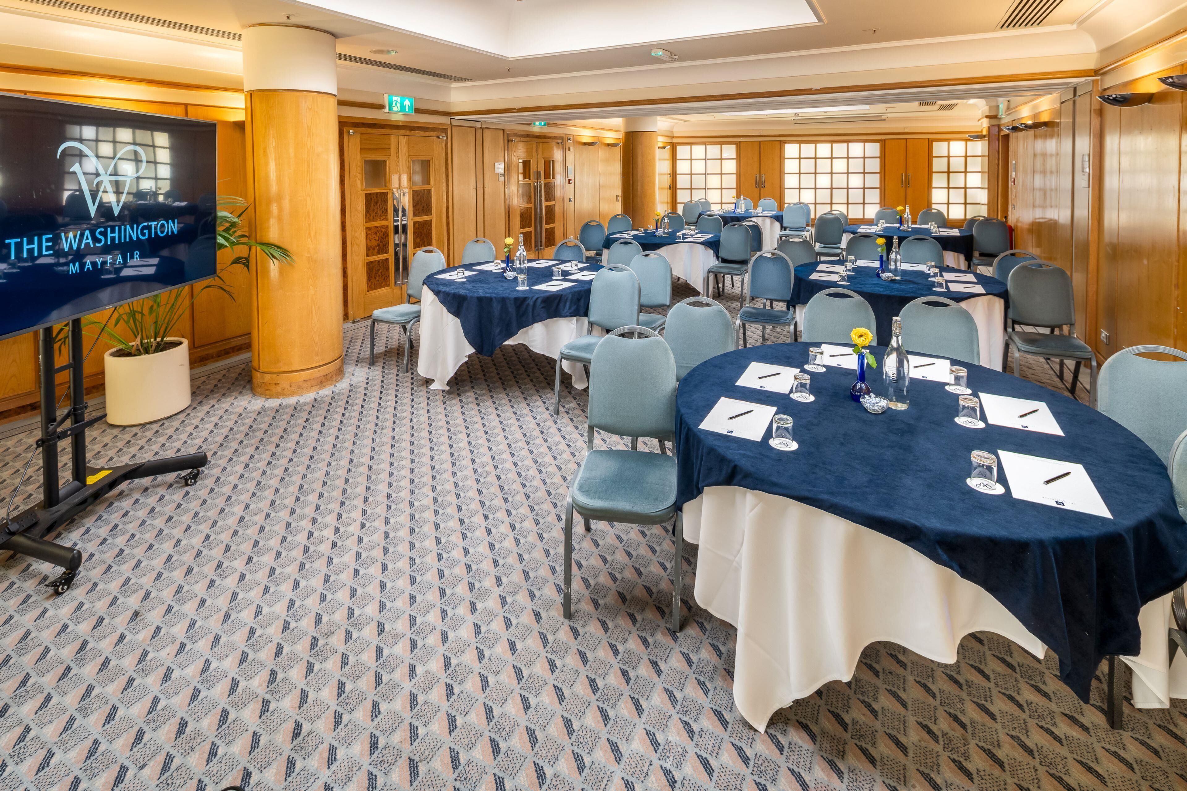 A conference room with round tables, chairs, and a large screen displaying 'THE WASHINGTON MAYFAIR'.