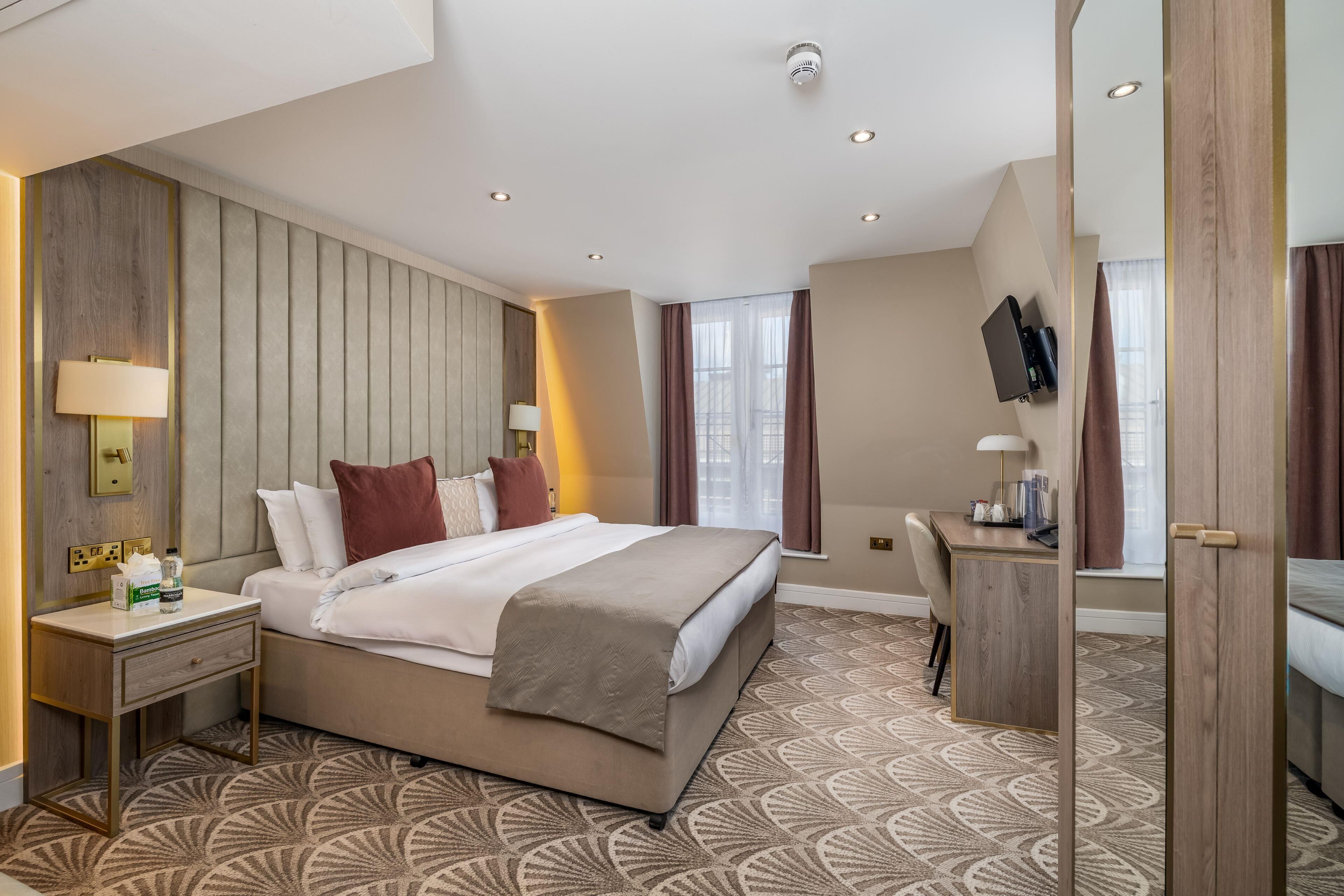 A hotel room with a bed, bedside table, lamp, TV, and a desk with a chair.