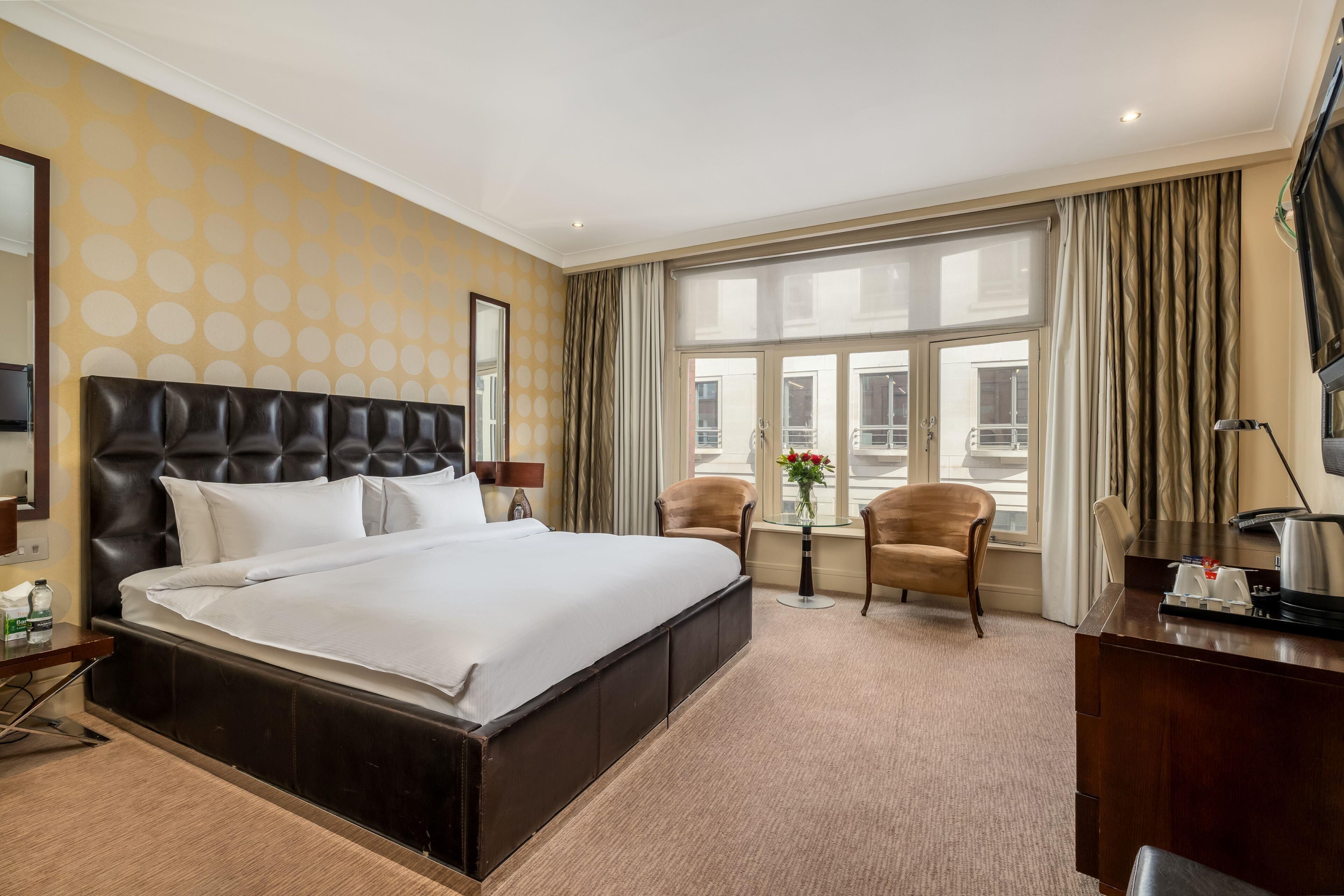 Une chambre d’hôtel avec un grand lit, un bureau, deux chaises, une petite table avec des fleurs et une grande fenêtre avec des rideaux.