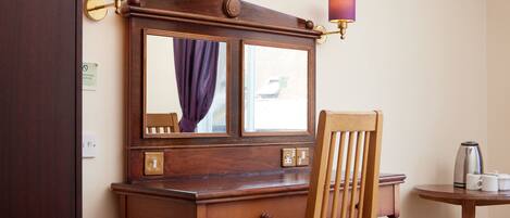 A wooden desk with a mirror, a chair, a television mounted above, and a small table with a kettle and cups.