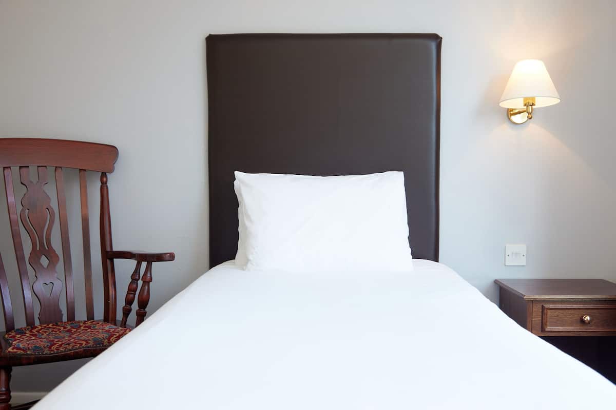 A neatly made bed with a white pillow and a dark headboard, a wooden chair with a patterned cushion, and a small bedside table with a lamp.