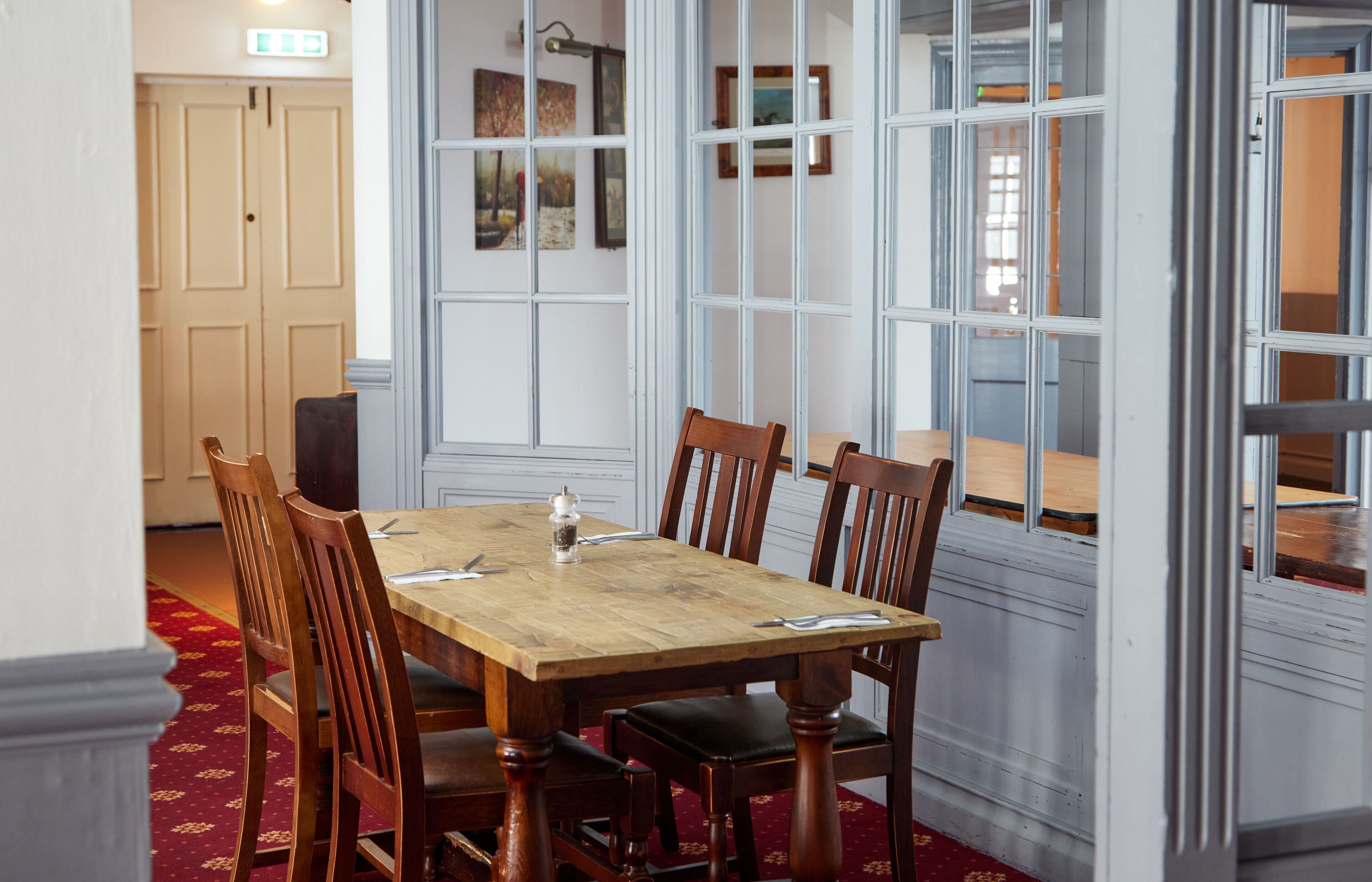 A dining area with a wooden table and chairs, a red carpet, and a view through large windows.