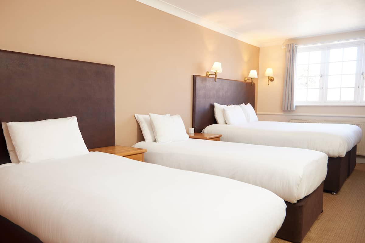 A hotel room with three single beds, each with white linens and a wooden headboard. There are bedside tables with lamps and a window with curtains.
