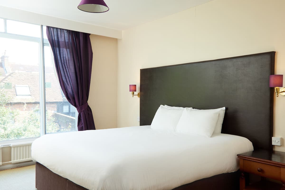 A hotel room with a large bed, a window with curtains, and two wall-mounted lamps.