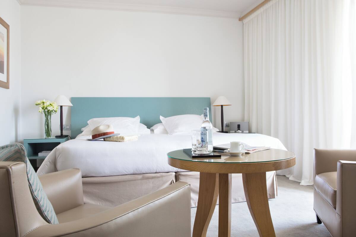 A hotel room with a bed, a round glass table, a sofa, and a nightstand with a lamp and a vase of flowers.