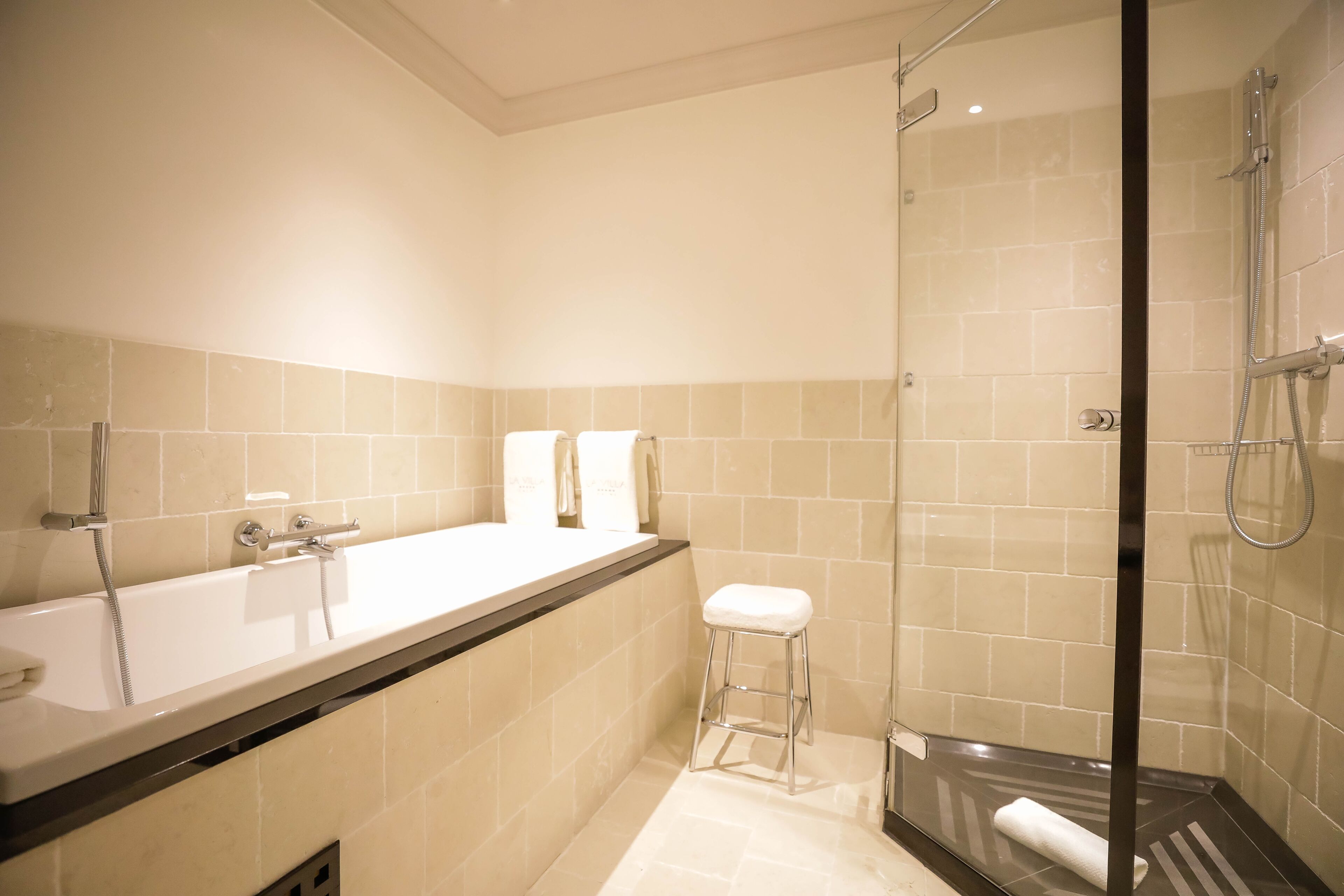 A modern bathroom with a glass-enclosed shower, a white bathtub, and a small stool.