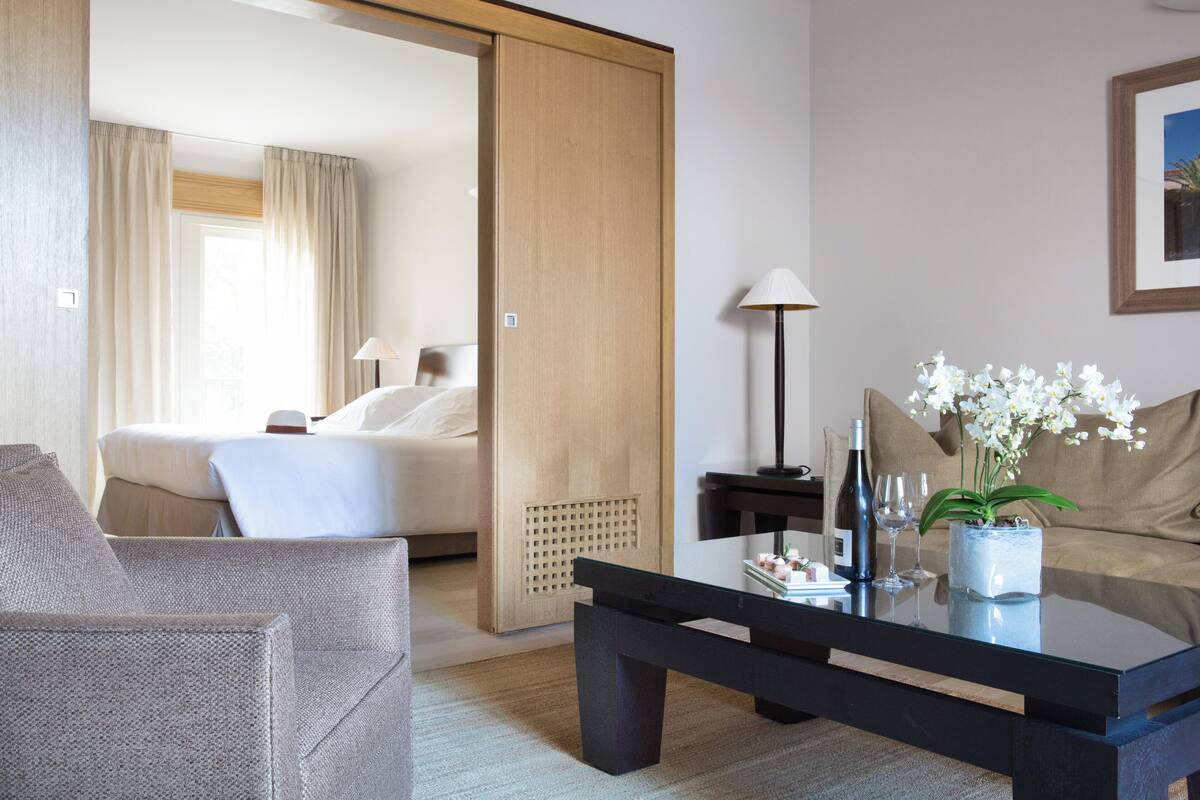 A hotel room with a bed, a sitting area with a sofa and armchair, a coffee table with a vase of flowers, and a lamp.