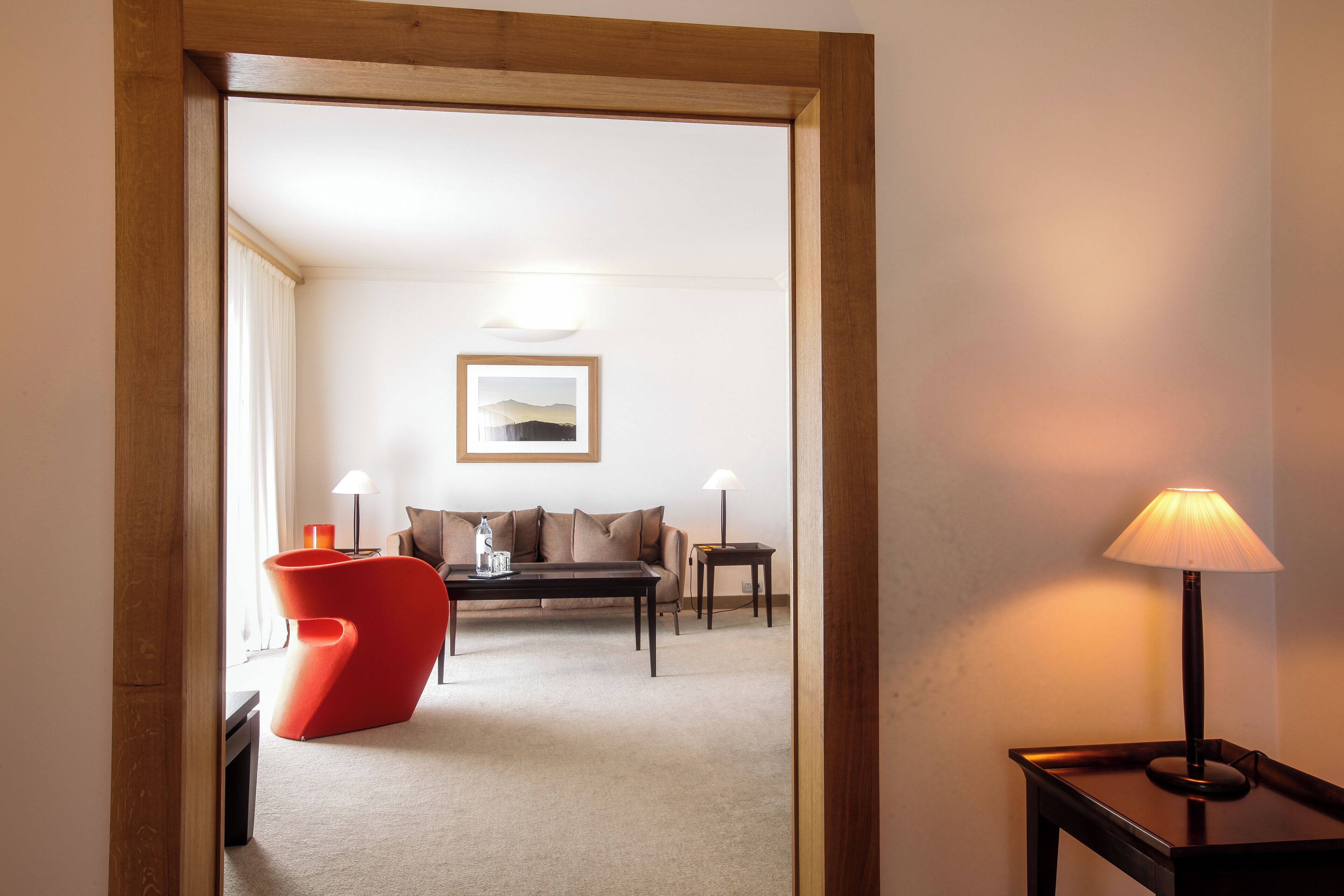 A hotel room with a modern red chair, a sofa, a small table, and a lamp.