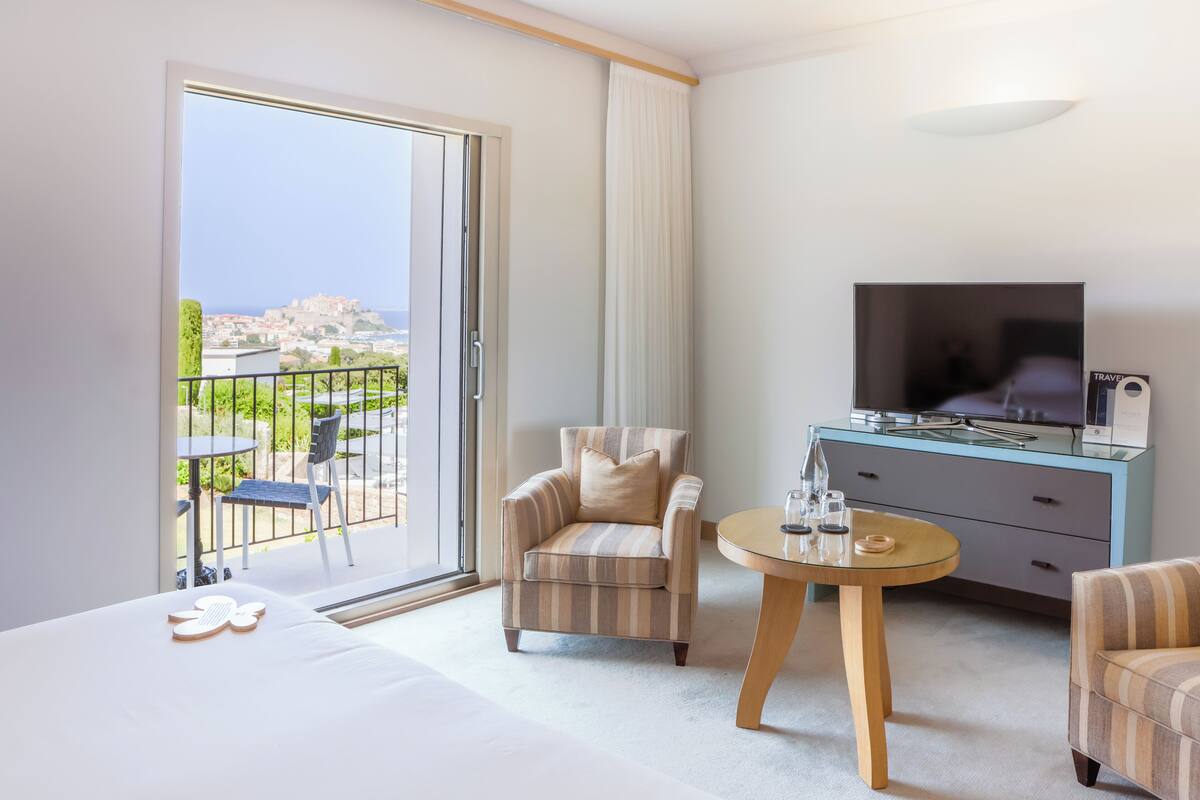 A hotel room with a balcony, a TV, a dining table, and two armchairs.