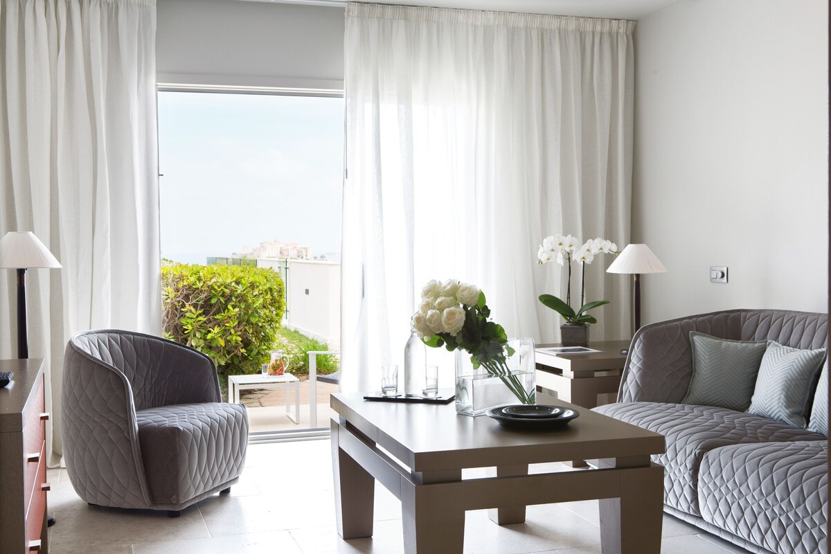 Un salon moderne avec un canapé, un fauteuil et une table basse. On y trouve une plante en pot et un vase rempli de fleurs sur la table.