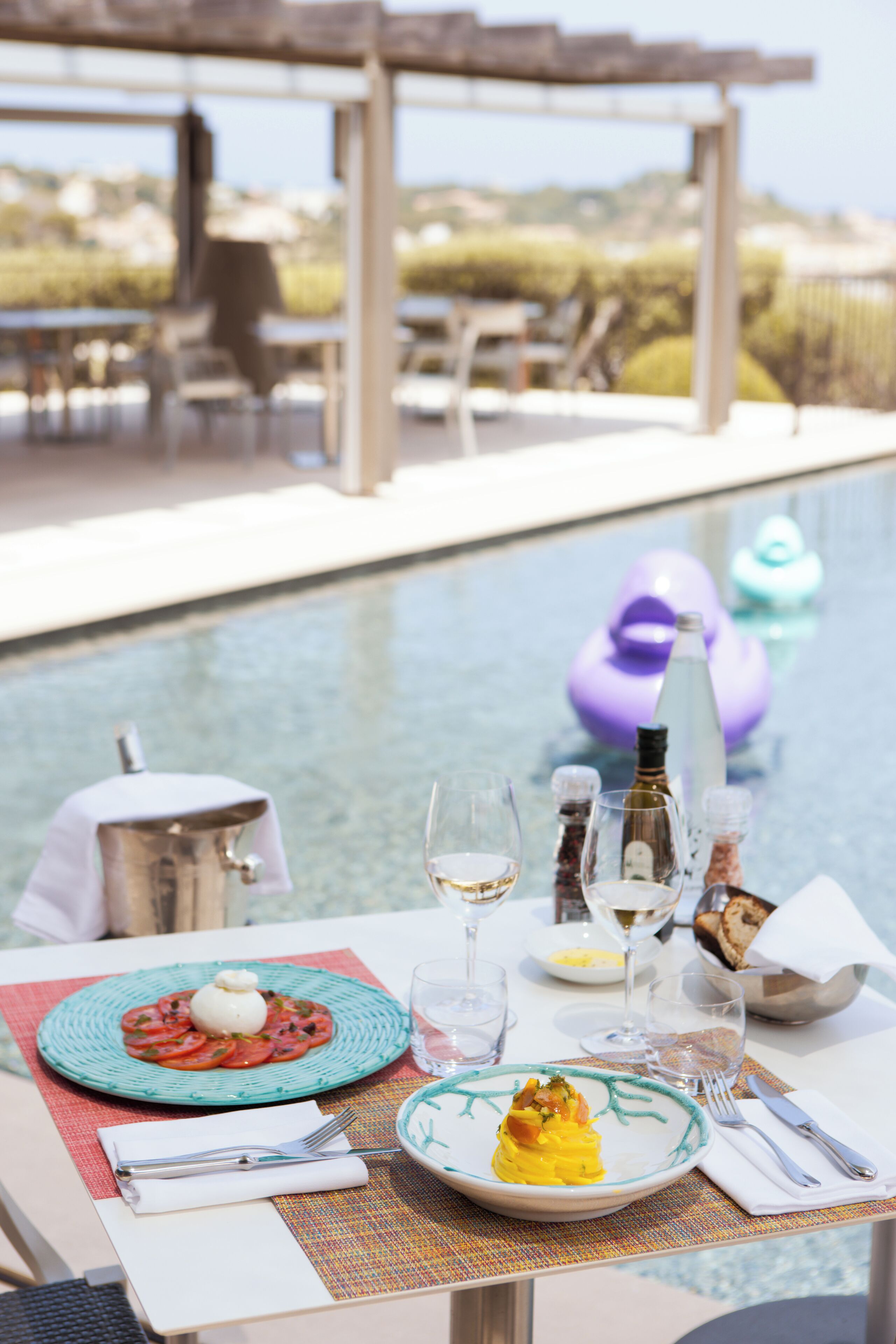 Une table à manger avec des assiettes remplies de nourriture, des verres à vin et une bouteille d’eau, le tout placé près d’une piscine.