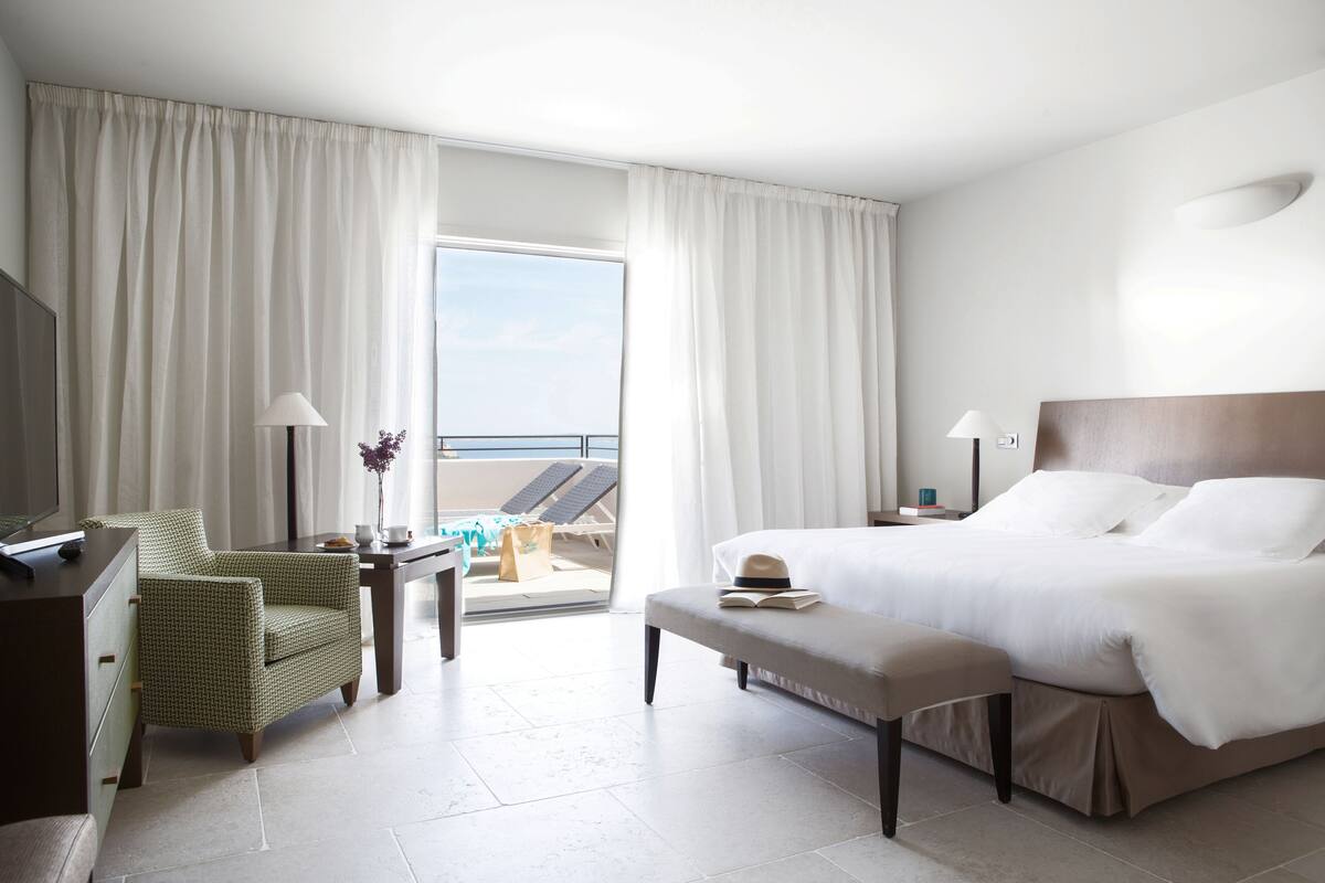 A hotel room with a bed, a desk, a chair, a TV, and a balcony with a view of the beach.