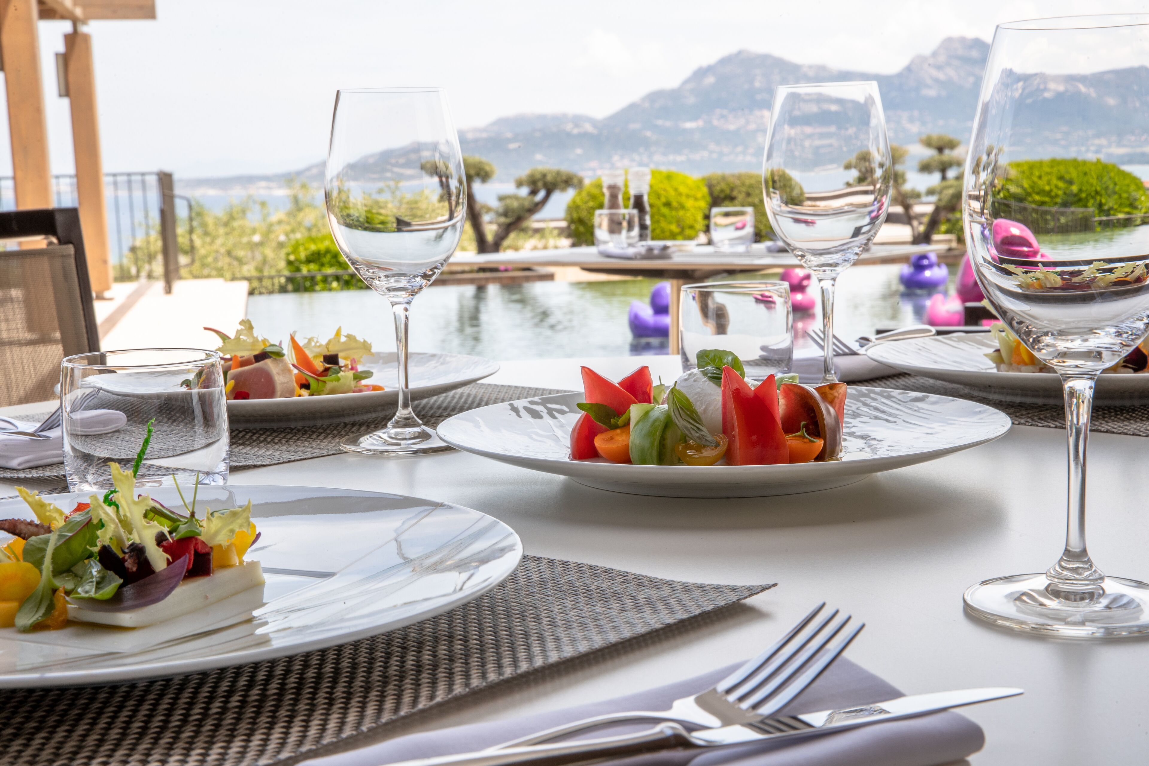 A dining table set with plates of food, wine glasses, and cutlery, overlooking a scenic landscape with mountains and water.