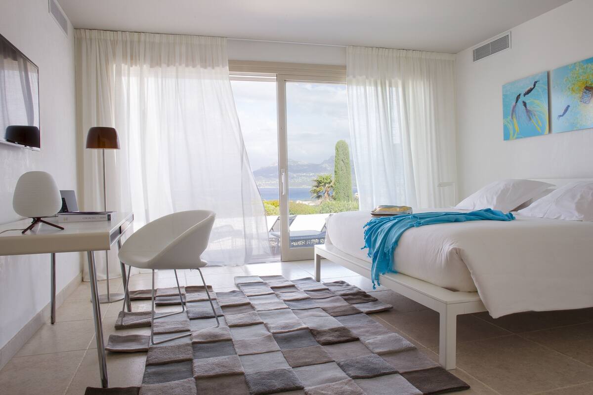 A hotel room with a bed, a desk with a chair, a view of the sea, and a painting on the wall.