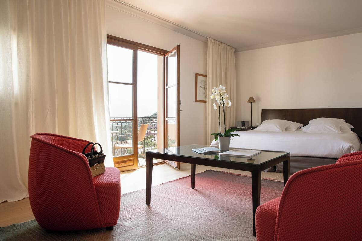 A hotel room with a bed, a desk, two red chairs, a small table with a vase of flowers, and a view of the outdoors through a large window.