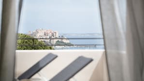 A balcony with two lounge chairs, a hat, and a view of a coastal town with buildings and a body of water. - La Villa Calvi (Calvi)