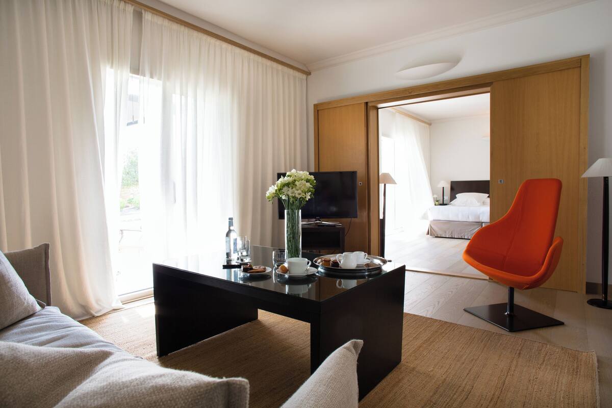 A modern hotel room with a sofa, a coffee table with a vase, a television, and an orange chair.