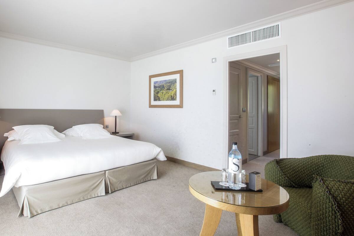 A hotel room with a bed, bedside tables, a lamp, a framed picture on the wall, a green chair, and a small round table with a bottle and glasses.