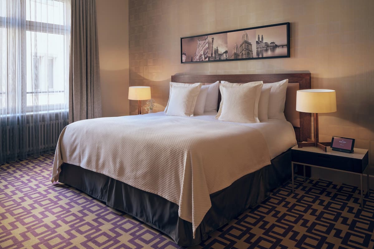Ein Hotelzimmer mit einem großen Bett, zwei Nachttischen mit Lampen, einem gemusterten Teppich und einem Stadtbild an der Wand.