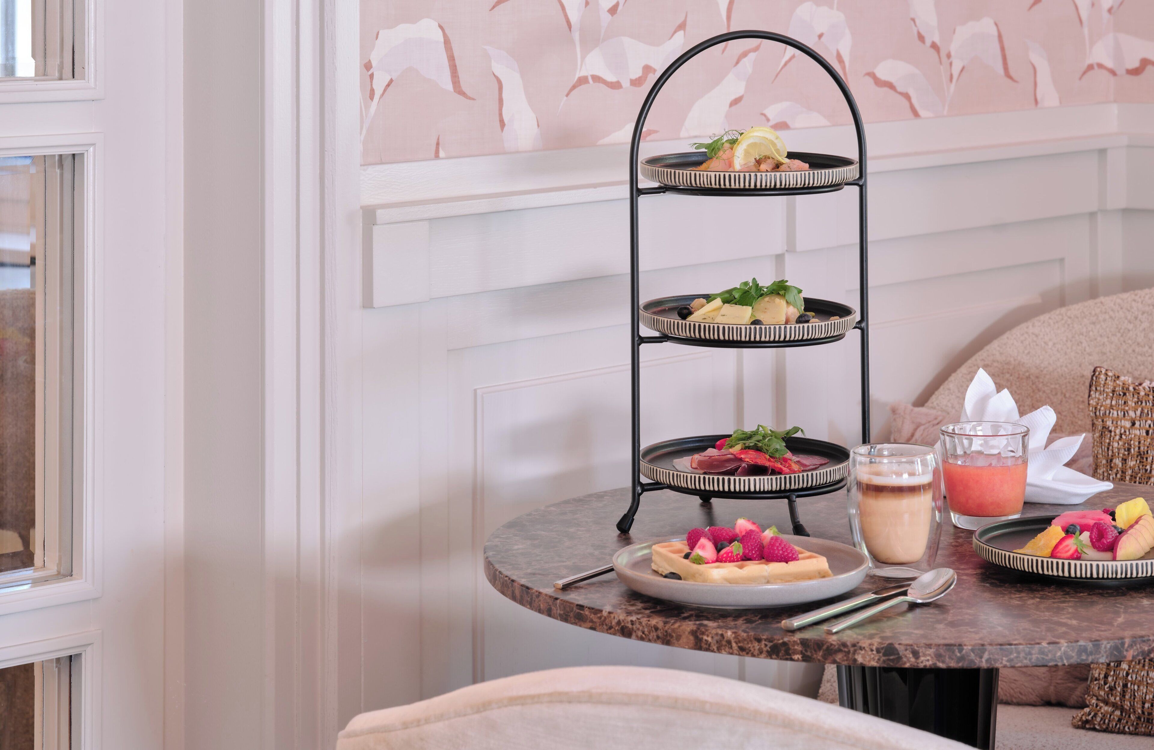 A three-tiered serving tray with food and drinks on a marble table, set against a floral wallpaper background.