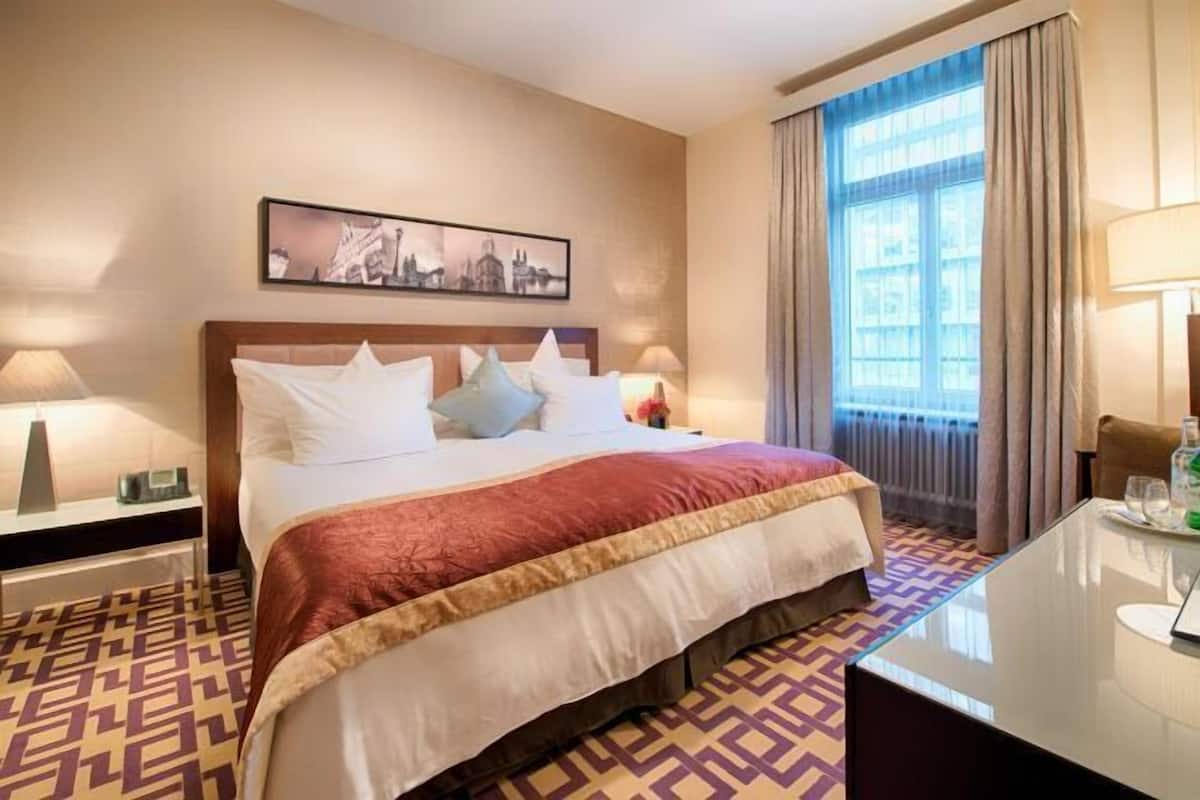 Ein Hotelzimmer mit Bett, Nachttisch, Lampe, einem Fenster mit Vorhängen und einem Stadtbild an der Wand.