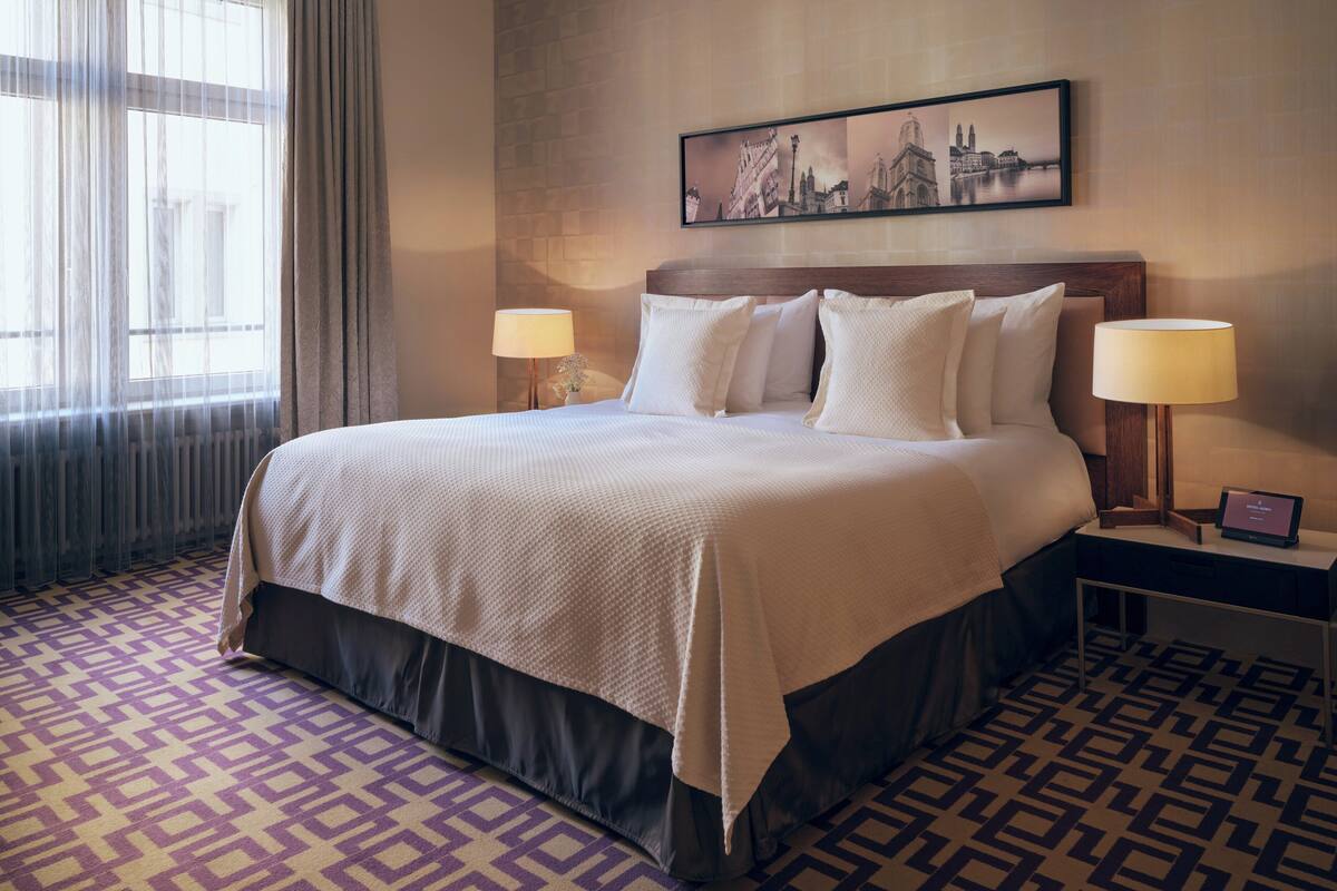 Ein Hotelzimmer mit einem großen Bett, zwei Nachttischen mit Lampen, einem gemusterten Teppich und einem Stadtbild an der Wand.