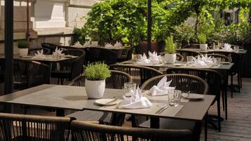 Un área de comedor al aire libre con mesas dispuestas para dos, rodeada de sillas de mimbre y plantas en macetas.