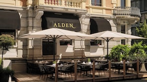 An outdoor seating area with tables and chairs under umbrellas, a building facade with the name 'ALDEN' displayed, and a brick wall. - Hotel Alden Splügenschloss Zürich - Leonardo Limited Edition (Zürich)