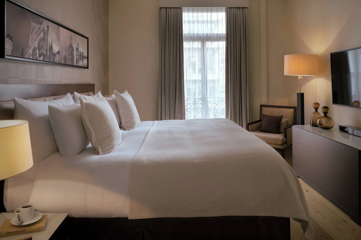Ein Hotelzimmer mit einem großen Bett, einem Nachttisch mit Lampe, einem Sessel, einer Kommode und einem an der Wand befestigten Fernseher.