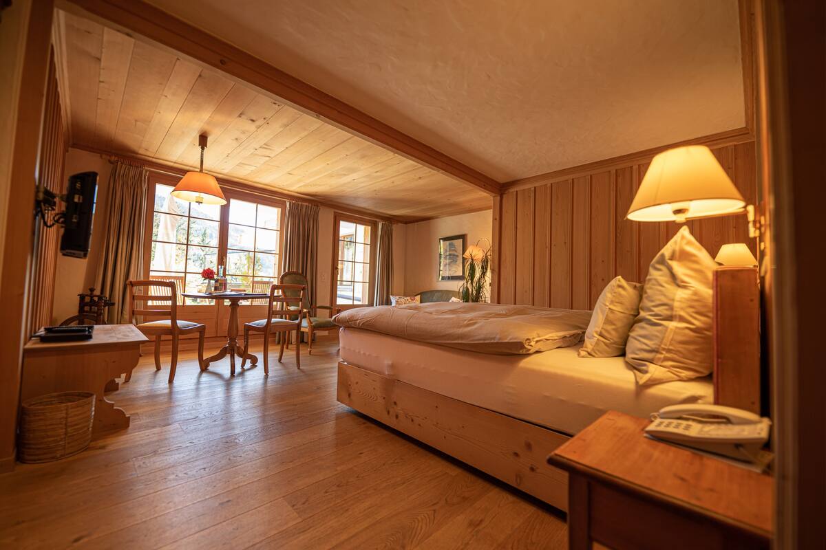 A cozy room with a bed, a desk, and a chair. There is a window with curtains, a lamp, and a telephone on the bedside table.