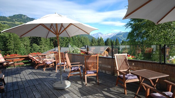 Une terrasse en bois, agréée de mobilier d’extérieur comprenant des chaises et une table, protégée par de grands parasols et offrant une vue sur un paysage montagneux.