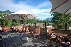 Une terrasse en bois, agréée de mobilier d’extérieur comprenant des chaises et une table, protégée par de grands parasols et offrant une vue sur un paysage montagneux.
