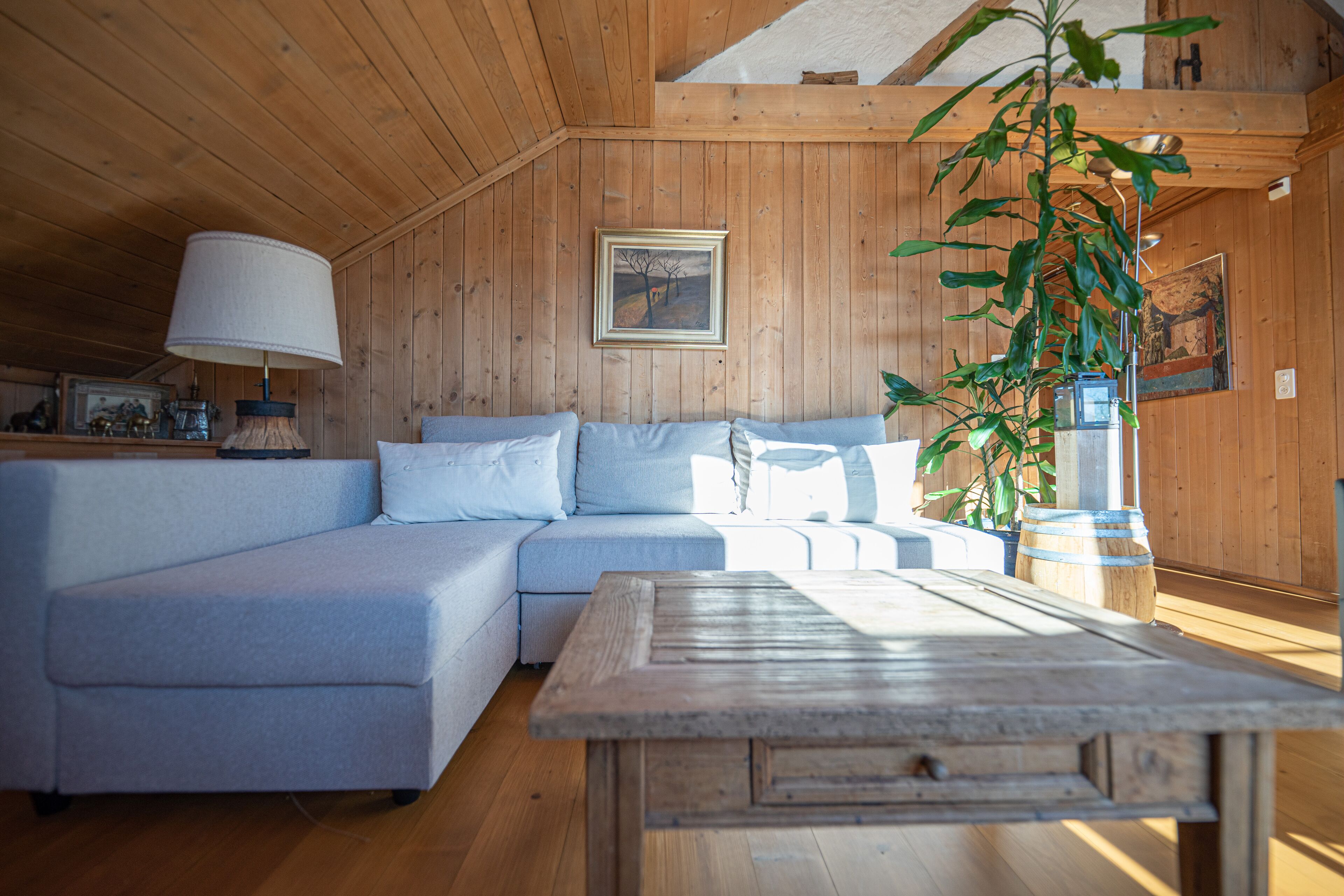 A cozy room with a wooden ceiling, a light blue sofa, a wooden table, and a large window allowing natural light.