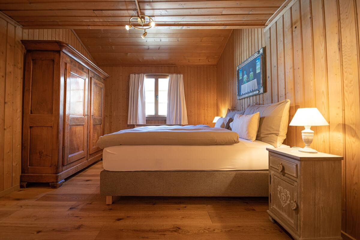 A wooden-paneled bedroom with a large bed, a nightstand, and a painting on the wall.