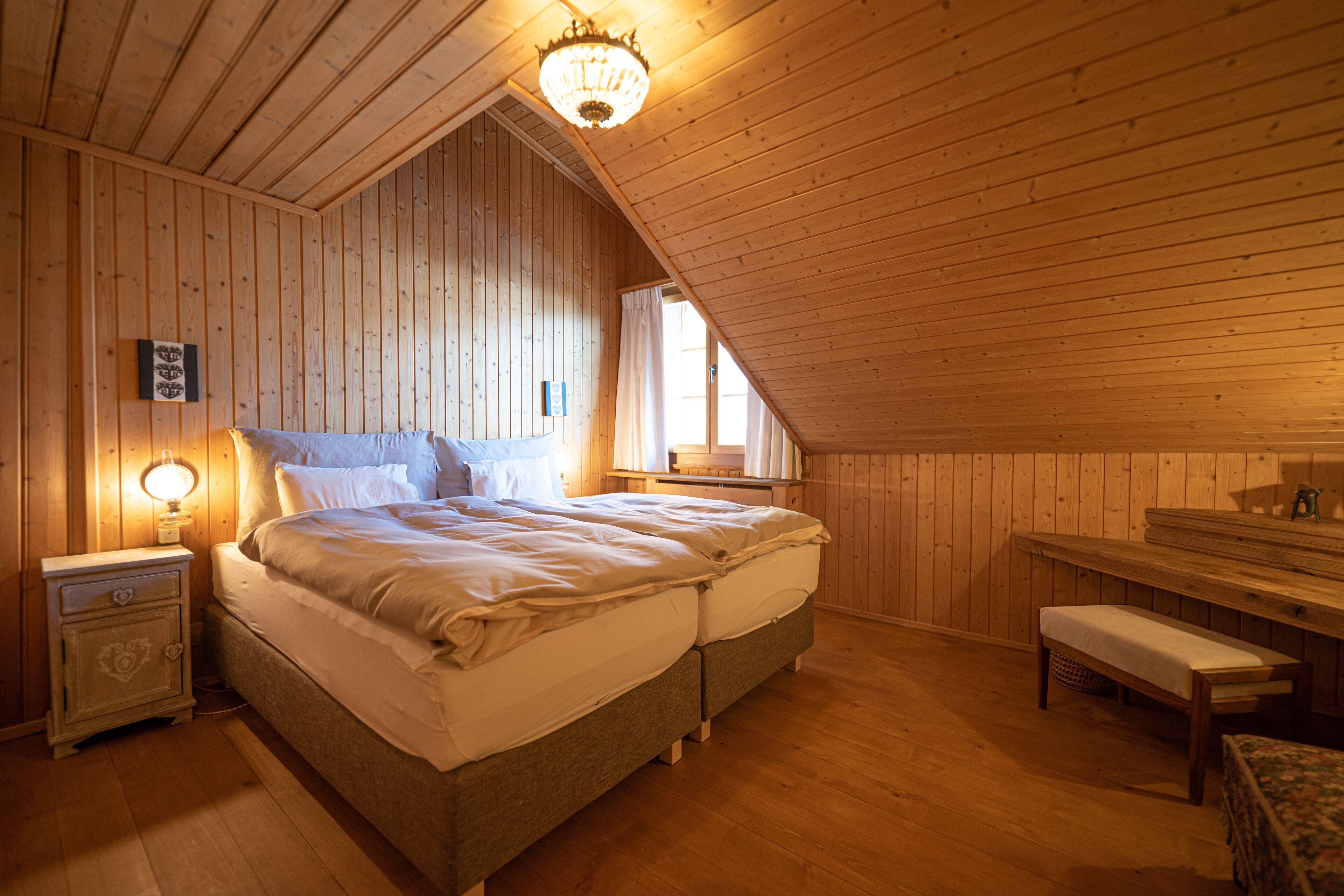 A cozy attic bedroom with a large bed, a nightstand, a bench, and a wooden ceiling.