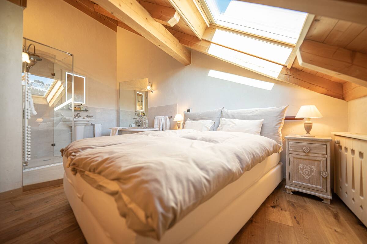 A bedroom with a large bed, skylight, wooden beams, a bedside table with a lamp, and a bathroom with a glass shower enclosure.