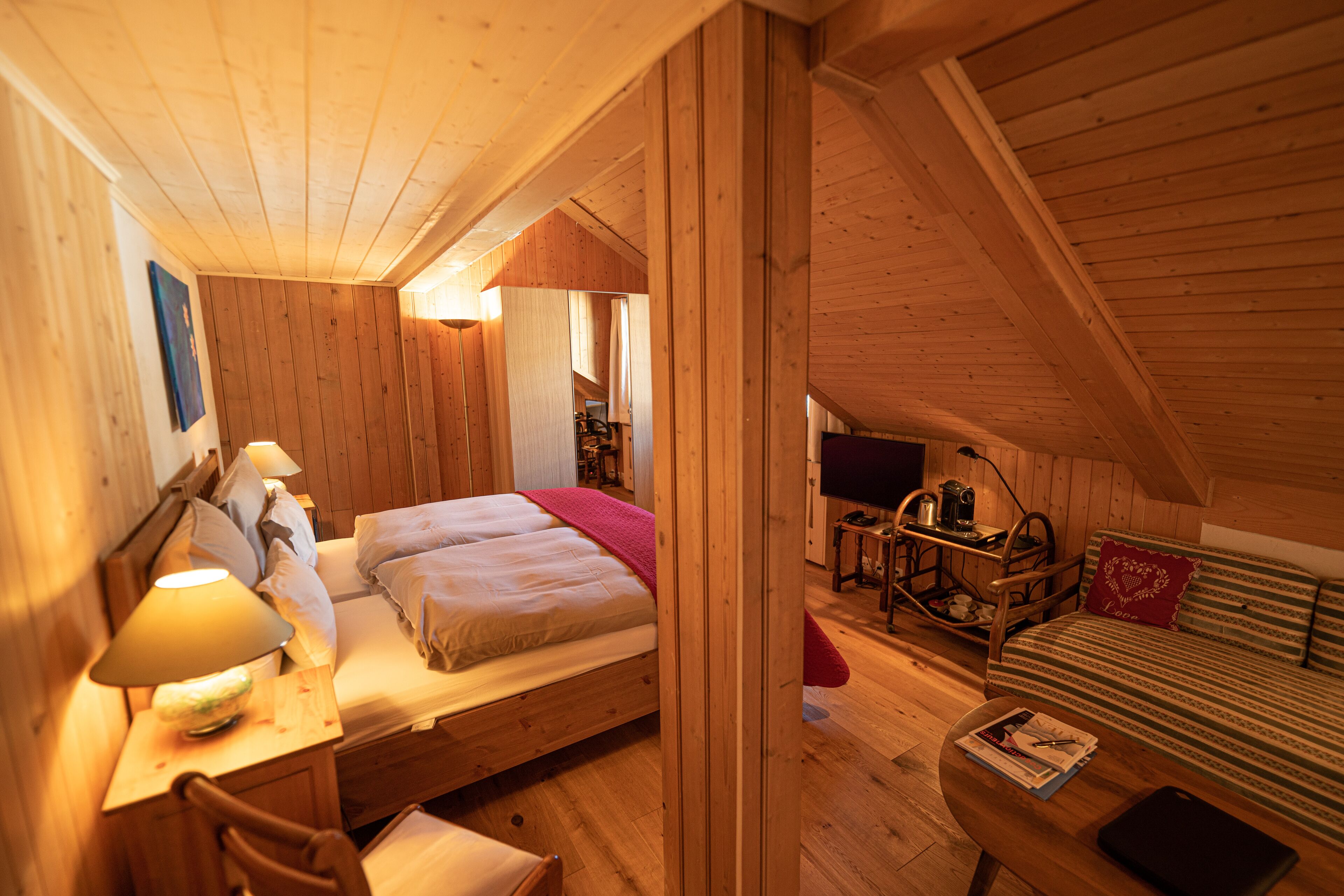 A cozy wooden cabin room with a bed, a desk with a lamp, a sofa, and a television.