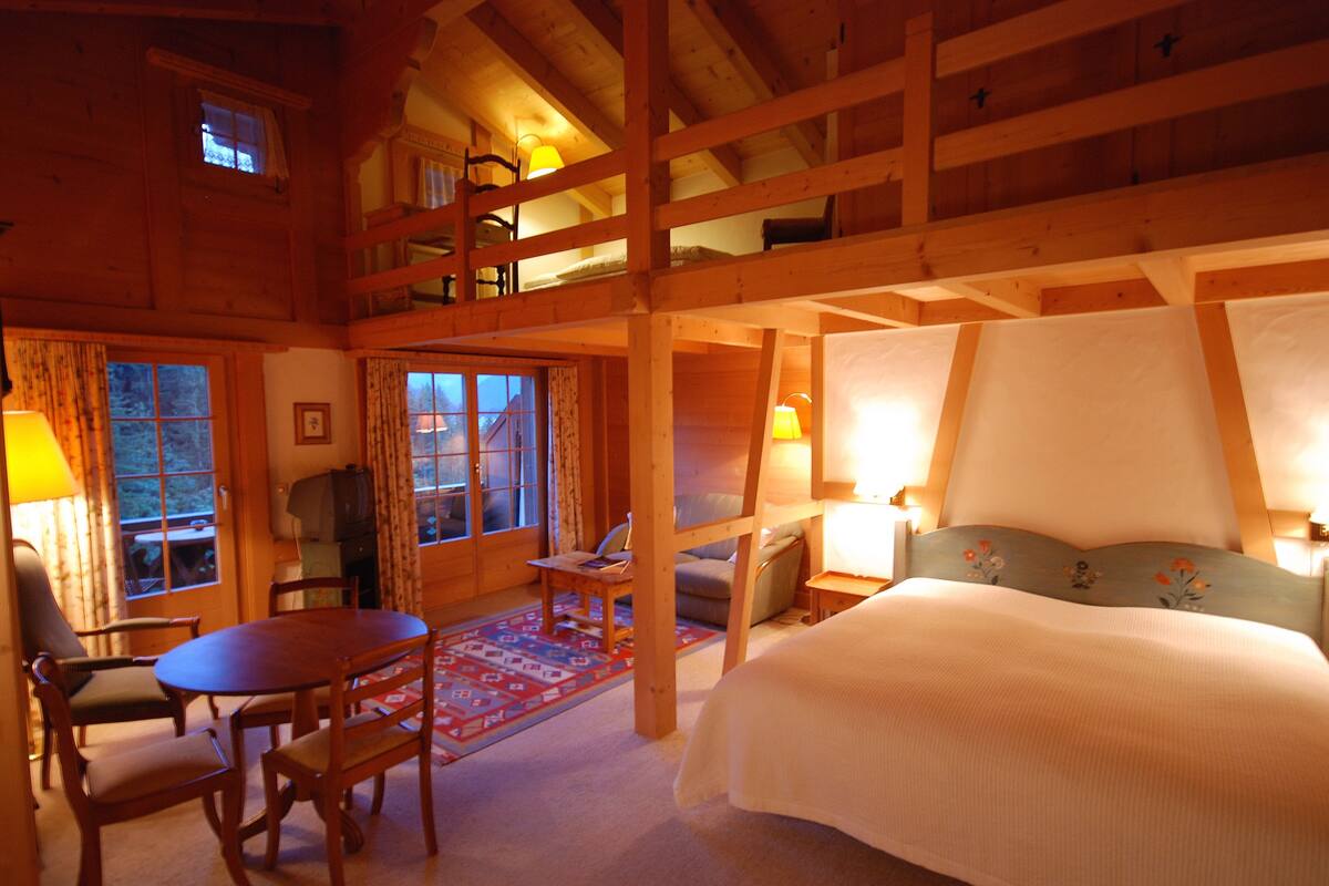 A cozy, wooden cabin-style room with a large bed, a dining table with chairs, a small table with a lamp, and a sofa.