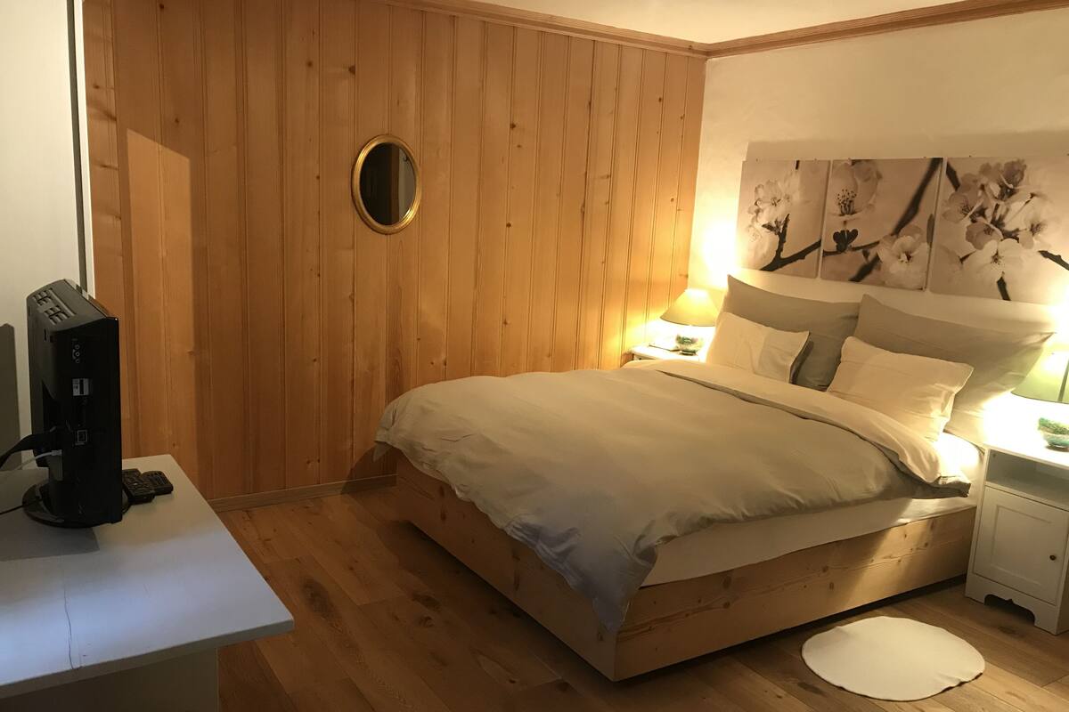 A bedroom with a wooden floor, a bed with white bedding, a desk with a computer, and a wall-mounted mirror.