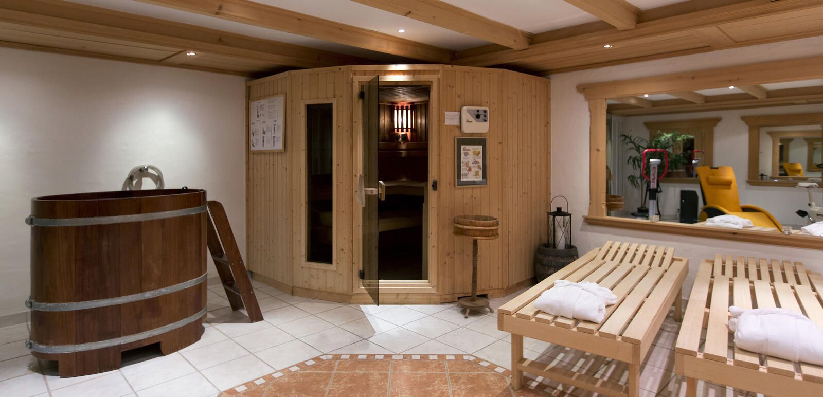 A sauna room with wooden benches, a wooden barrel, and a mirror reflecting a yellow chair.