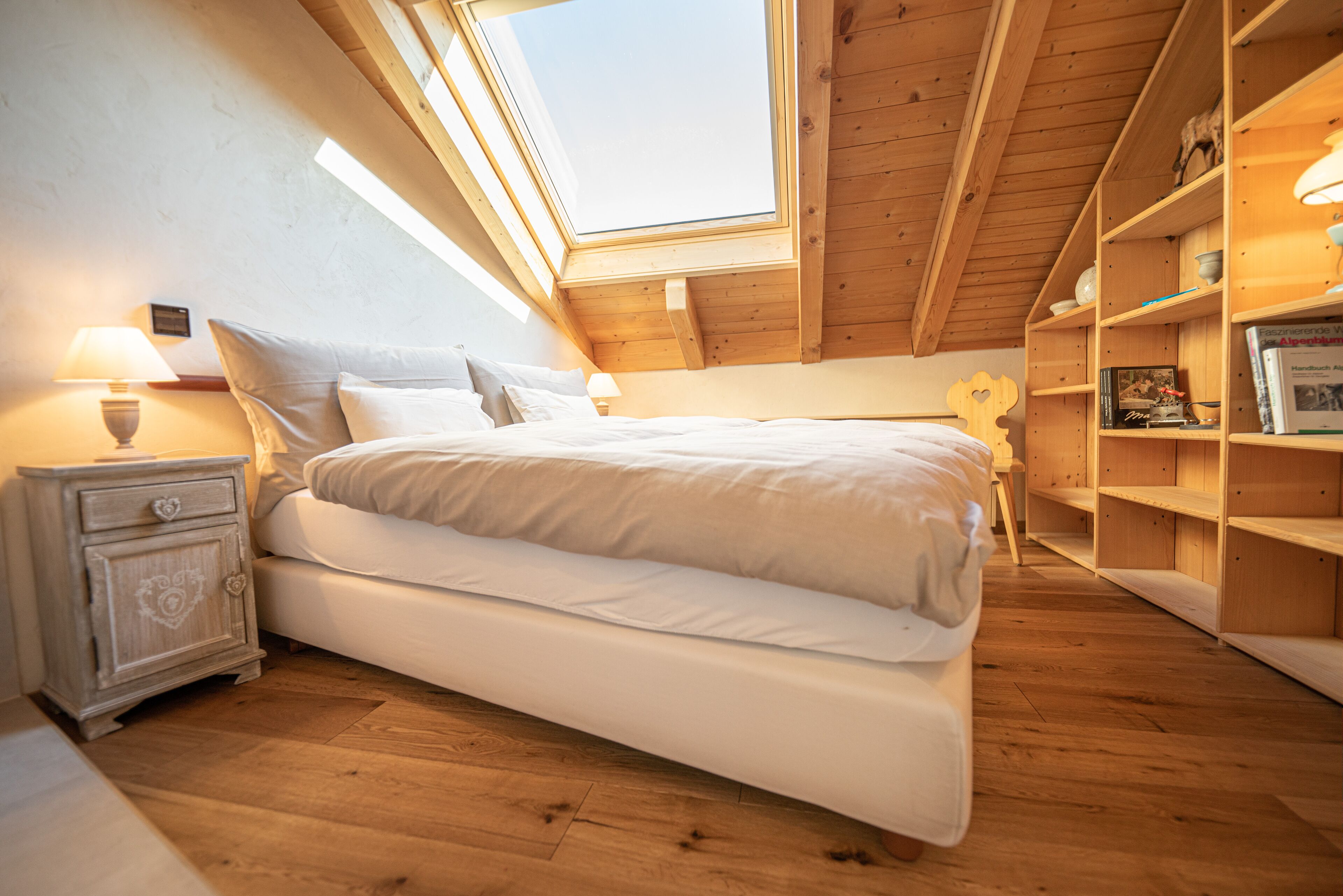 A bedroom with a skylight, wooden floor, a bed with white bedding, bedside tables with lamps, and a wooden bookshelf.