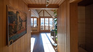 Un couloir en bois, avec une grande peinture sur le mur de gauche, une plante sur celui de droite, et une vue sur l’extérieur grâce à de grandes fenêtres.