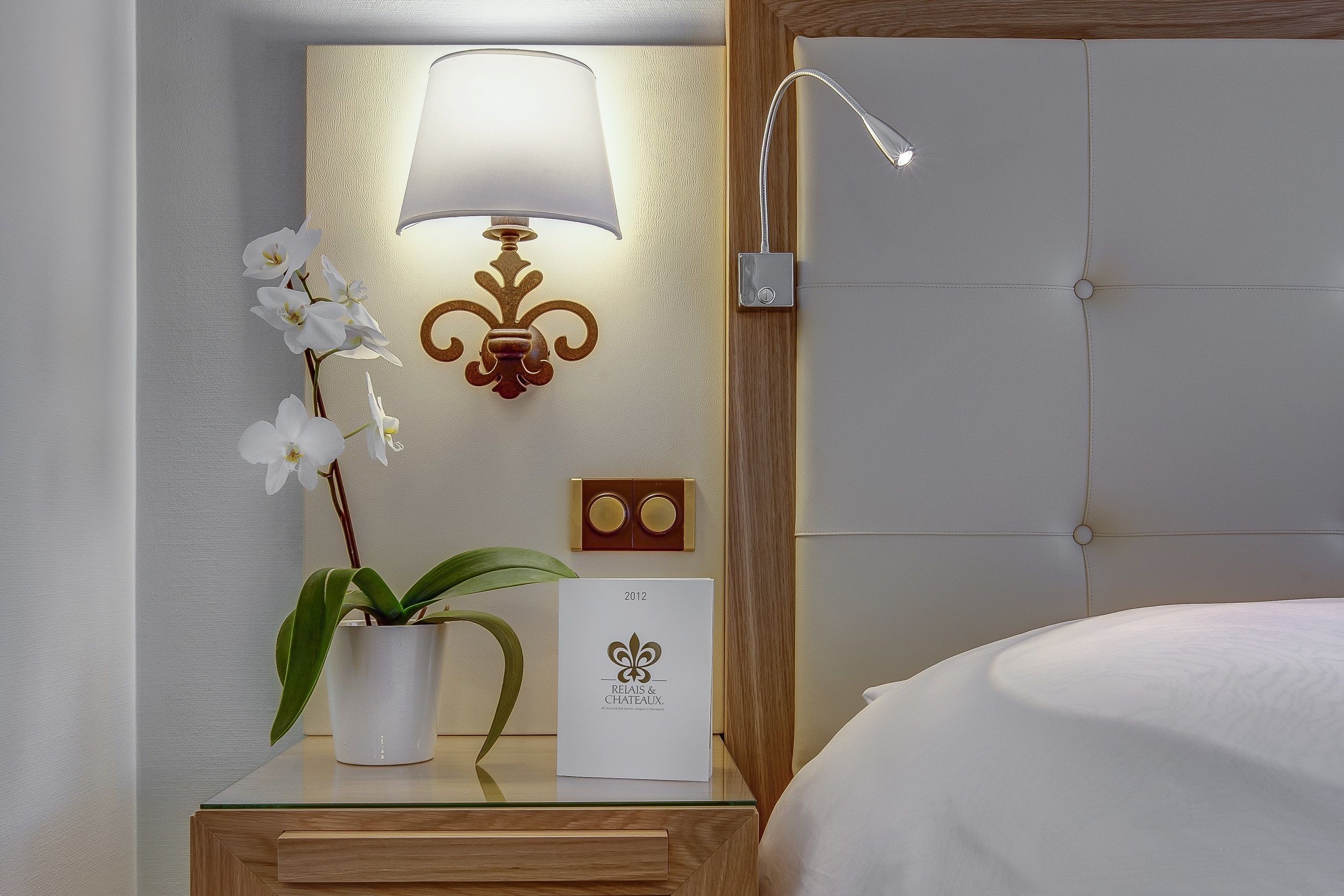 Une chambre d’hôtel bien rangée, avec une table de chevet, une lampe, une orchidée en pot et un certificat encadré.