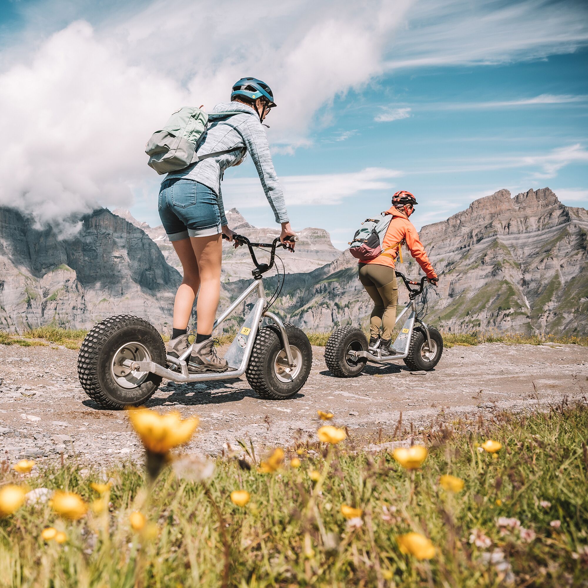 Two people riding electric bikes on a mountain trail with rocky terrain and mountains in the background.