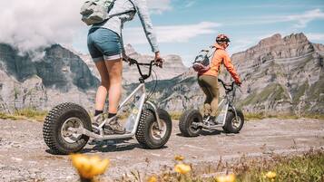 Two people riding electric bikes on a mountain trail with rocky terrain and mountains in the background.