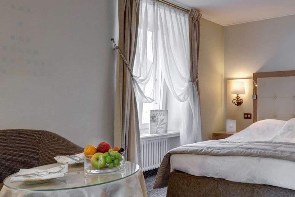 Une chambre d’hôtel avec un lit, une table en verre sur laquelle se trouvent des fruits, une chaise et une fenêtre avec des rideaux.