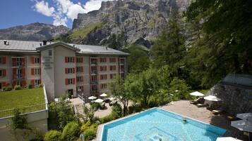 A hotel with a swimming pool surrounded by greenery and mountains in the background.