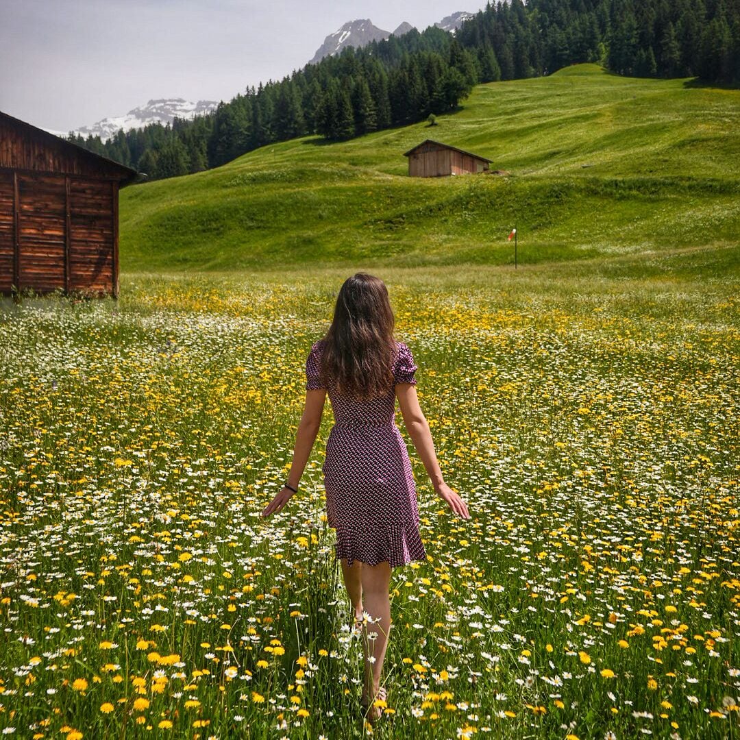 Una mujer caminando por un campo de margaritas, con una cabaña de madera y montañas al fondo.