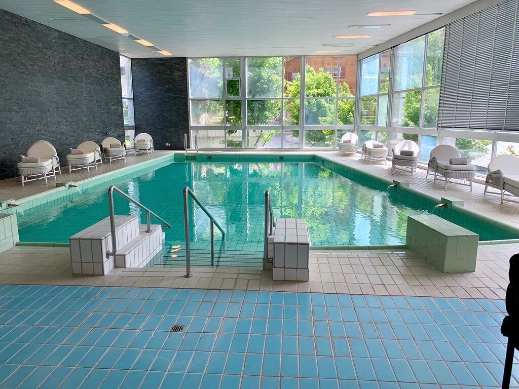 An indoor swimming pool with clear blue tiles, surrounded by white lounge chairs and a large window offering a view of greenery outside.