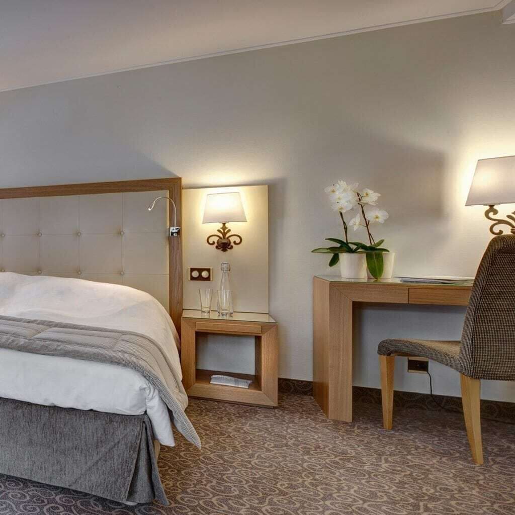 A hotel room with a bed, a desk, a chair, a nightstand, a lamp, and a potted orchid.